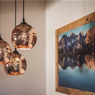 Drei hängende Lampen mit metallischem Schimmer und ein gerahmtes Bild von Bergen am Wasser. Die Atmosphäre ist modern und einladend.