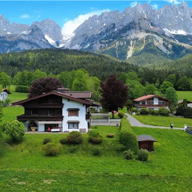 A picturesque landscape with a traditional house and majestic mountains in the background.  
Surrounded by green meadows and trees, the scene radiates tranquility and natural beauty.