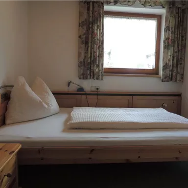A simple bedroom with a wooden bed and two decorative cushions. The room has a window with curtains and a wooden wardrobe.