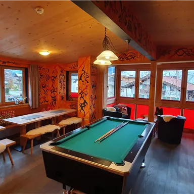 A cozy room with a pool table in the middle and large windows overlooking a beautiful view. The walls are colorfully decorated, and there is comfortable seating.
