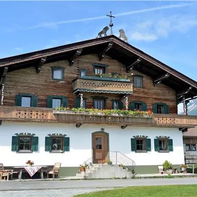 Ein traditionelles alpenländisches Haus mit Holzverkleidung und einem großen Balkon. Im Hintergrund sind Berge und eine grüne Wiese zu sehen.