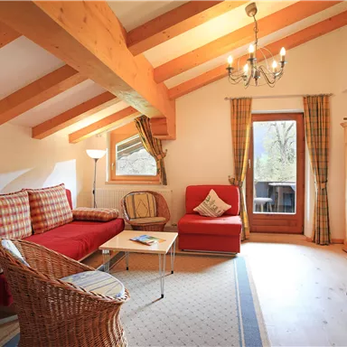 A cozy living room with wooden beams and a red couch. Large windows provide a view outside, and the light creates an inviting atmosphere.