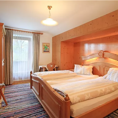 A cozy bedroom with two wooden beds and a bright decor. The room has large windows and a carpet in vibrant colors.
