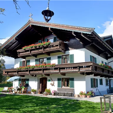 A traditional alpine house with a wide balcony and blooming flowers. It stands in a green setting under a clear blue sky.