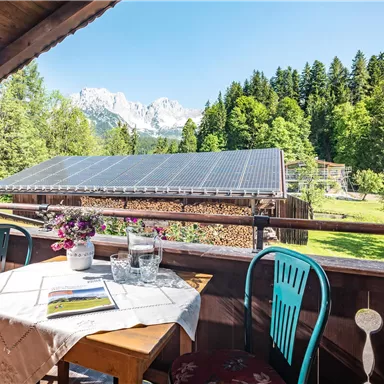A cozy balcony with a table and chairs, surrounded by trees and mountains. In the background, solar panels can be seen under a clear blue sky.