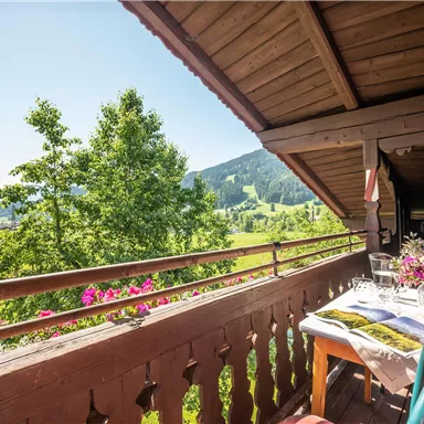A cozy balcony with a table and a flower view of a green landscape. In the background, gentle hills and a blue sky can be seen.
