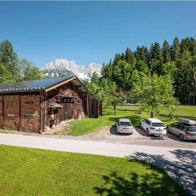A rustic building surrounded by green meadows and trees. Snow-capped mountains can be seen in the background.