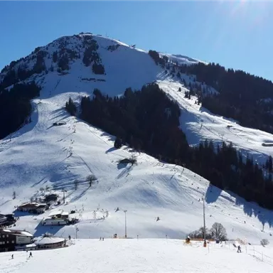 Eine schneebedeckte Berglandschaft mit mehreren Skipisten. Im Vordergrund sind kleine holzverkleidete Gebäude zu sehen.