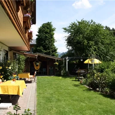 A beautiful garden with a well-maintained lawn and many plants. Next to the house, there are tables with yellow tablecloths and a sun umbrella.