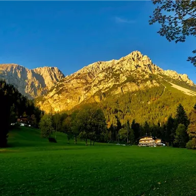 An impressive mountain landscape with high peaks and a clear blue sky. In the foreground, a green meadow and some trees can be seen.