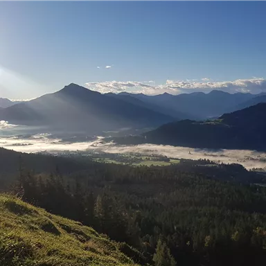 Eine malerische Berglandschaft mit Nebel im Tal und Sonnenstrahlen, die durch die Wolken scheinen. Die Szenerie vermittelt ein Gefühl von Ruhe und Schönheit der Natur.
