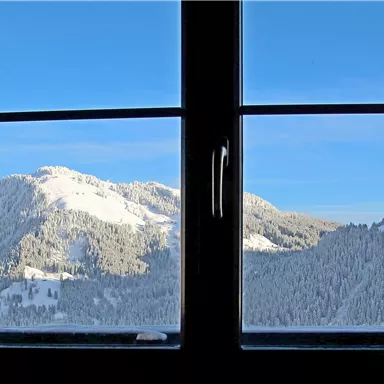 Blick aus dem Fenster auf schneebedeckte Berge unter einem klaren blauen Himmel. Die Landschaft ist ruhig und winterlich.