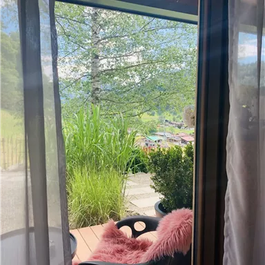 Ein schöner Ausblick von einem Fenster auf eine grüne Landschaft mit Pflanzen. Auf der Terrasse steht ein bequemer Stuhl mit einem pinken Kissen.