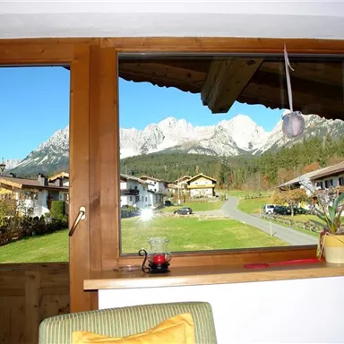 Blick aus einem Fenster auf eine malerische Berglandschaft mit klar blauem Himmel. Im Vordergrund sind traditionelle Häuser und eine grüne Wiese zu sehen.
