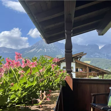A picturesque balcony with colorful flowers and a view of majestic mountains. The sky is clear and the landscape is green and inviting.