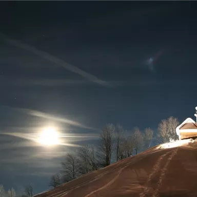 Eine verschneite Landschaft bei Nacht mit einem hellen Mond. Im Vordergrund steht ein kleines Gebäude auf einem Hügel, umgeben von Bäumen.
