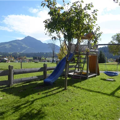 Ein Spielplatz mit einer Rutsche und einer Schaukel in einer grünen Wiese. Im Hintergrund sind Berge und ein blauer Himmel zu sehen.