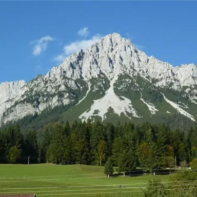A majestic mountain with an impressive rock structure. In the foreground, green meadows and forests stretch out.