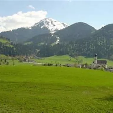 Eine malerische Landschaft mit grünen Wiesen und schneebedeckten Bergen im Hintergrund. Im Vordergrund steht eine kleine Kirche.