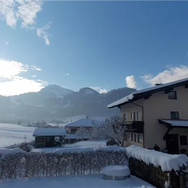 Ein Winterlandschaft mit schneebedeckten Häusern und Bergen im Hintergrund. Der Himmel ist blau mit wenigen Wolken.