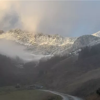 Eine mystische Berglandschaft mit schneebedeckten Gipfeln und nebelverhüllten Hängen. Im Vordergrund ist ein kurvenreicher Weg zu sehen.