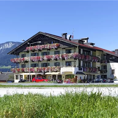 A cozy hotel with many flowers on the balcony. In the background, mountains and a clear blue sky can be seen.
