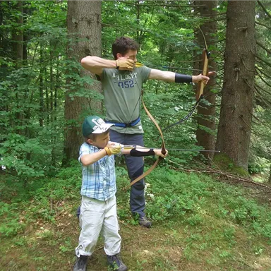 An adult and a child are practicing archery in the woods. They are standing in a green environment among tall trees.