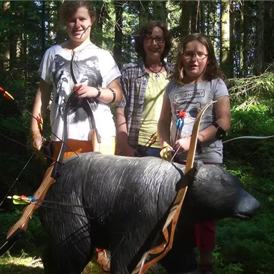 Three people are standing in the forest next to a carved bear figure with arrows. It is an active environment with plenty of greenery and sunlight.