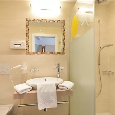 Ein modernes Badezimmer mit einem Waschbecken und einer Dusche. Die Wände sind hell und schlicht gestaltet.
