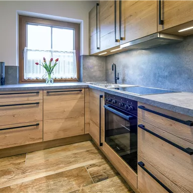 Eine moderne Küche mit Holzschränken und einem grauen Arbeitsbereich. Auf der Fensterbank stehen frische Blumen in einer Vase.