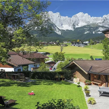 Ein malerischer Garten mit Liegen, umgeben von traditionellen Häusern und majestätischen Bergen im Hintergrund. Der Himmel ist klar und strahlend blau.