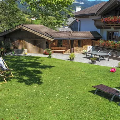Ein ruhiger Garten mit Liegen und Kunstwerken. Im Hintergrund ist ein gemütliches Holzhaus und eine zwei-stöckige Fassade zu sehen.