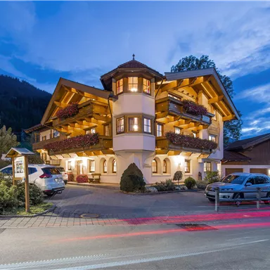 Ein hübsches Gebäude im alpinen Stil mit mehreren Balkonen. Die Umgebung ist von Bäumen umgeben und dämmernd beleuchtet.
