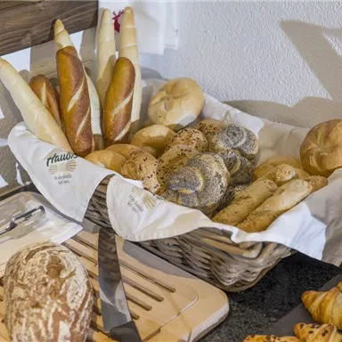 A basket with various types of bread, including baguettes, rolls, and croissants. The breads are carefully arranged and covered with a white cloth.