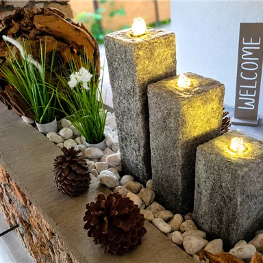 Decorative area with three illuminated stone columns and plants. A welcome sign and pine cones enhance the cozy atmosphere.