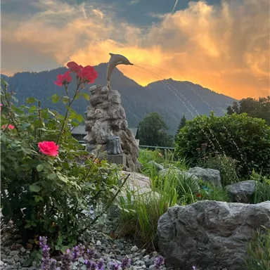 A beautiful garden with red roses and purple flowers. In the background, majestic mountains rise beneath a colorful sky.