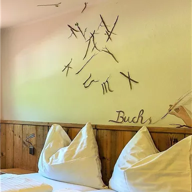 A cozy bedroom with two pillows and wood paneling. On the wall, there are artistic branches and the lettering "Buchs" visible.