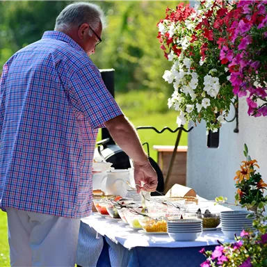 A man is standing at a table with various foods outdoors. In the background, colorful flowers and a sunny environment can be seen.