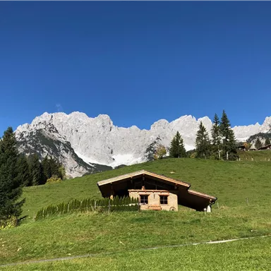 A picturesque mountainous region with a traditional wooden house. In the background, tall, rocky mountains rise under a clear blue sky.