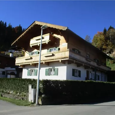 Ein traditionelles Holzhaus in den Bergen, umgeben von Bäumen. Die Fassade hat grüne Fensterläden und einen Balkon.
