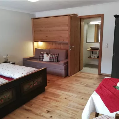 Ein gemütliches Zimmer mit zwei Betten und einem Sofa. Der Boden ist aus Holz und es gibt einen kleinen Tisch mit einer dekorativen Tischdecke.