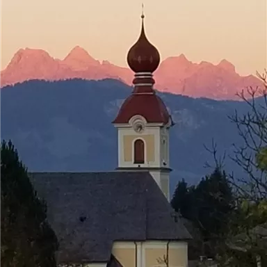 A church with a distinctive dome and a red roof stands in front of majestic mountains. The sky is illuminated in soft colors.