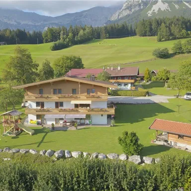 Eine malerische Landschaft mit mehreren Gebäuden und grünen Wiesen. Im Hintergrund sind Berge und ein klarer Himmel zu sehen.