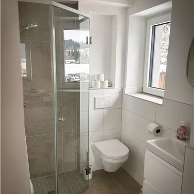 A modern bathroom with a glass shower and a white toilet. There is a window that lets in light and a small sink cabinet.