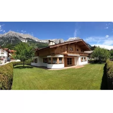 A charming wooden house with a well-maintained garden and green trees. In the background, majestic mountains rise under a clear sky.