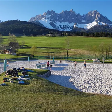 Ein Sandplatz für Volleyball in einer malerischen Umgebung mit hohen Bergen im Hintergrund. Die Sonne scheint auf eine Gruppe von Menschen, die aktiv spielen.
