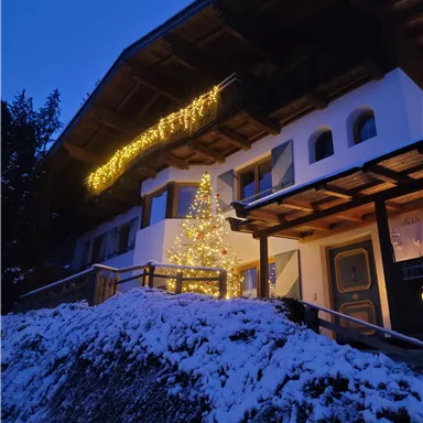 Ein gemütliches Haus im Winter mit Schnee und festlicher Beleuchtung. Ein Weihnachtsbaum steht vor der Eingangstür und sorgt für eine einladende Atmosphäre.