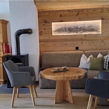 Eine gemütliche Wohnzimmersituation mit einem Holztisch, zwei Stühlen und einer Couch. Der Raum hat eine warme Holzverkleidung und eine gemütliche Atmosphäre.