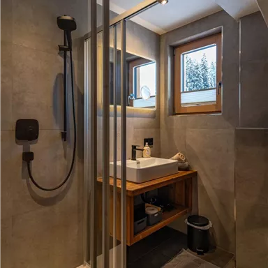 A modern bathroom with a glass shower and wooden shelf. Daylight streams in through the window.
