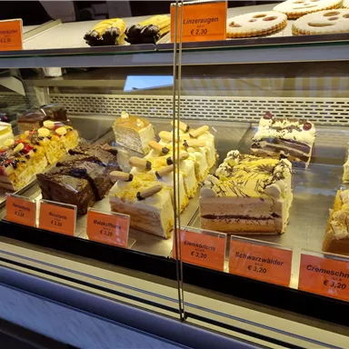 Eine Vitrine mit verschiedenen Torten und Kuchen. Die Leckereien sind bunt dekoriert und mit Preisen versehen.